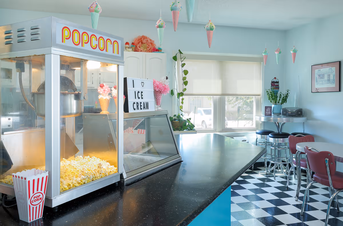 Interior of a retro-style dining area with a popcorn machine on the counter, a sign that says 'I ♥ ICE CREAM', hanging ice cream cone decorations, checkered black and white floor, red chairs, and a small round table with stools near a window.