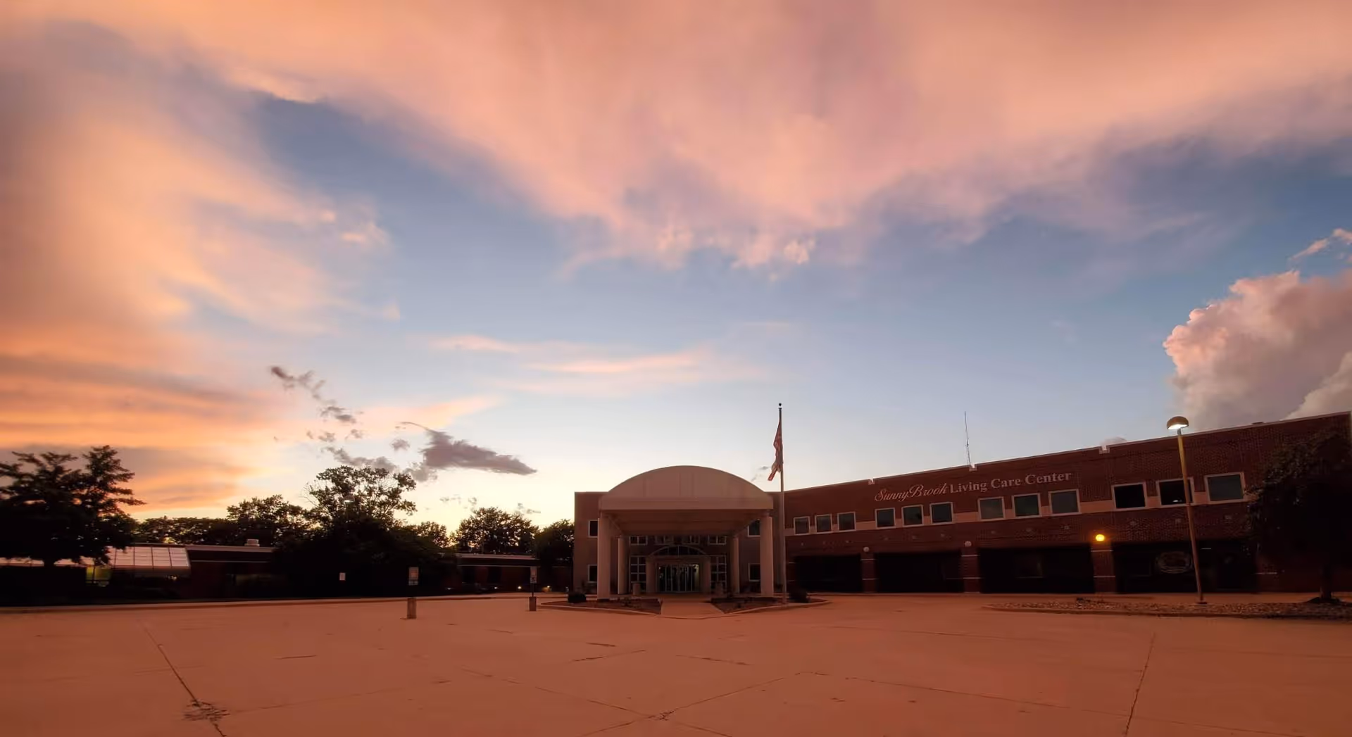 Exterior view of a large brick building with an entrance covered by a white canopy and an American flag flying in front. The sky is colorful with a sunset showing pink and orange clouds. Trees surround the building and there is a large empty paved area in front.