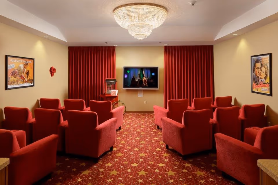 A small theater room with red upholstered armchairs arranged in rows facing a wall-mounted flat screen TV. The room has red curtains behind the TV, a popcorn machine to the left, and movie posters on the beige walls. The carpet is red with a star pattern, and a large chandelier hangs from the ceiling.