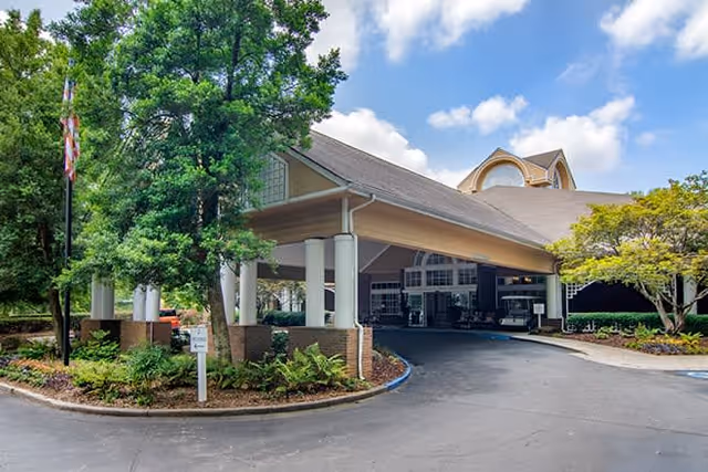 Entrance of a senior living facility with a covered driveway, surrounded by trees and landscaping under a partly cloudy sky.