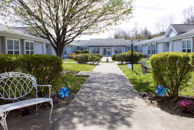 A sunny outdoor courtyard at The Addison of Uniontown featuring a concrete walkway lined with trimmed bushes and small blue pinwheel decorations. There is a white metal bench on the left side under a tree with green leaves. The courtyard is surrounded by single-story buildings with white siding and multiple windows.