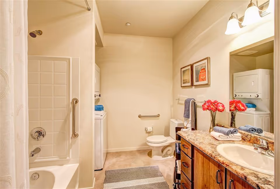 A bathroom with a bathtub and shower on the left, a toilet in the center, and a sink with a granite countertop and wooden cabinets on the right. Above the sink is a large mirror with two light fixtures. There are two framed pictures on the wall above the toilet, a towel rack with a blue towel, and a striped rug on the floor. A stacked washer and dryer unit is visible in the corner near the toilet. A vase with red flowers and neatly folded towels are on the countertop.