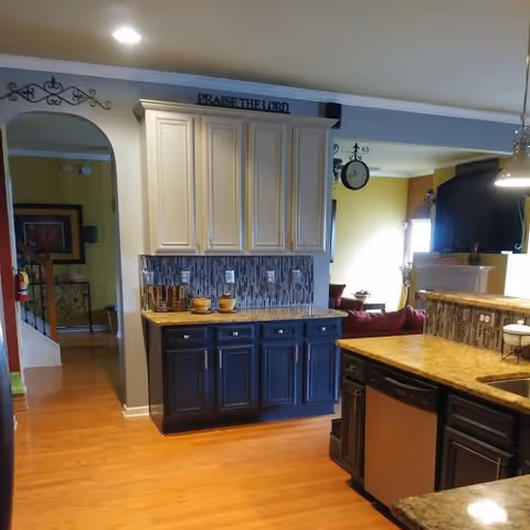 Interior view of a kitchen area with wooden floors, two-tone cabinets with dark lower cabinets and light upper cabinets, a granite countertop, a tiled backsplash, and a dishwasher. The kitchen opens into a living room area with a clock on the wall and a window letting in natural light. A decorative sign above the cabinets reads 'PRAISE THE LORD'.