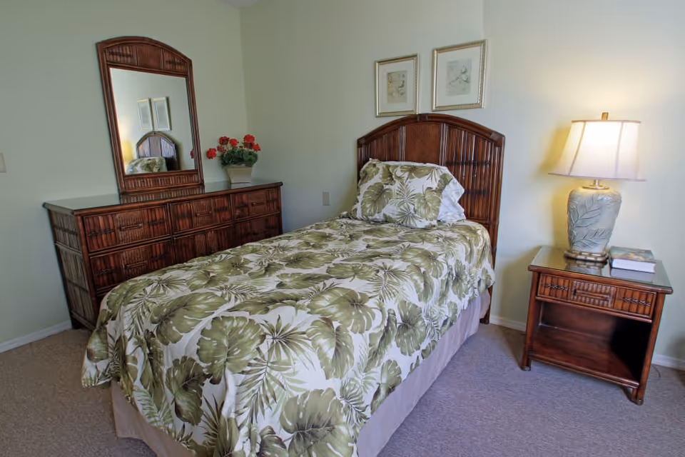 A bedroom with a single bed covered in a green and white leaf-patterned bedspread and matching pillow. Next to the bed is a wooden nightstand with a decorative lamp and two books. On the opposite side of the bed is a wooden dresser with a large mirror and a vase of red flowers. The walls are light-colored and there are two framed pictures hanging above the bed.