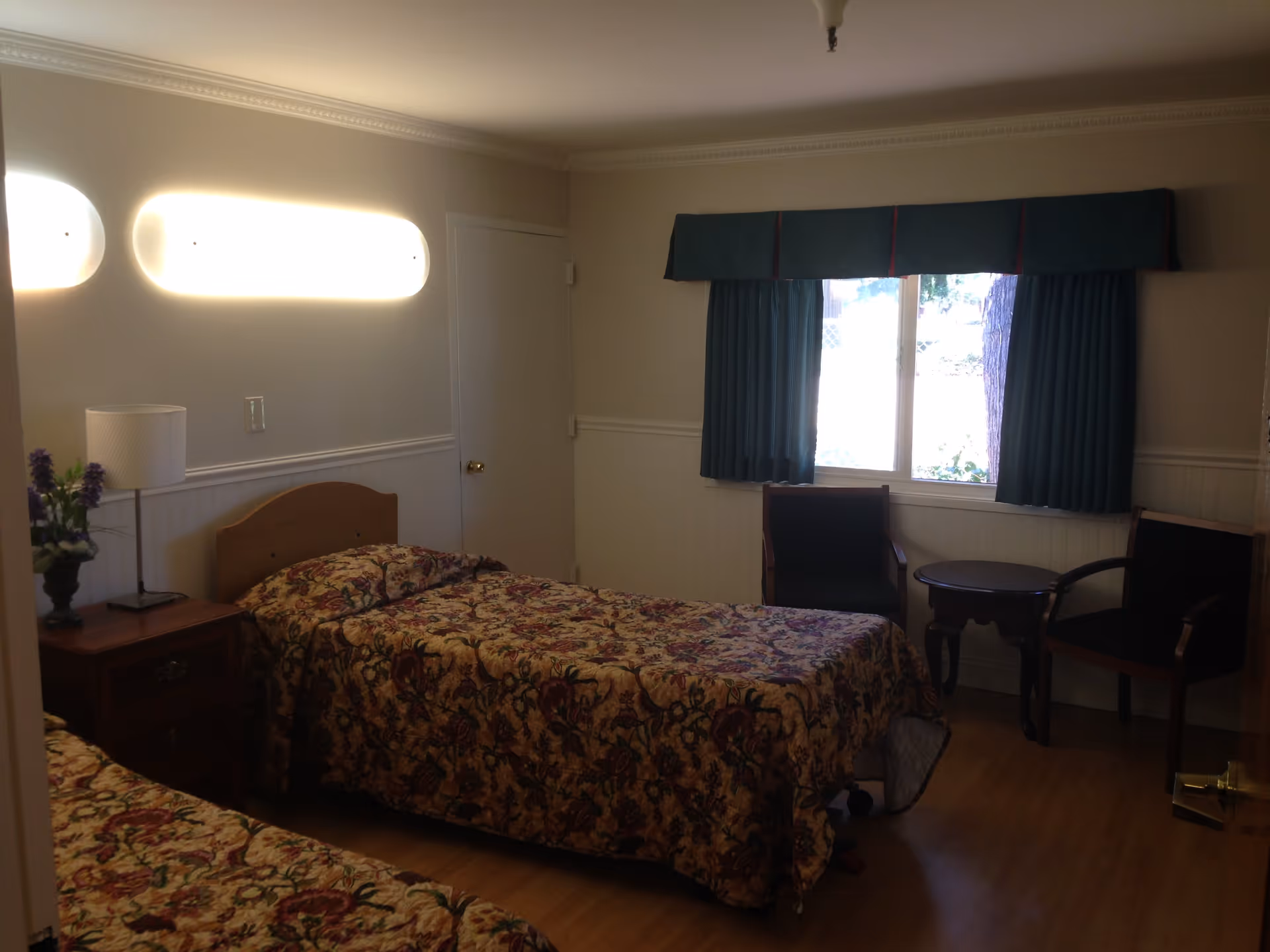 A small bedroom with two single beds covered in floral patterned bedspreads. There is a wooden nightstand with a lamp and a vase of flowers between the beds. A window with dark green curtains and a valance is on the far wall, with two wooden chairs and a small round table beneath it. The room has light-colored walls and wood flooring.
