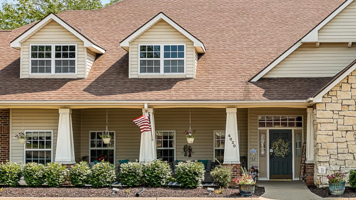 Front exterior view of a residential-style building with beige siding, a brown shingled roof, two dormer windows, a covered porch with white columns, hanging flower pots, an American flag, and a green front door with a wreath. The building number 4020 is visible on one of the columns, and there are shrubs and flower pots along the front.