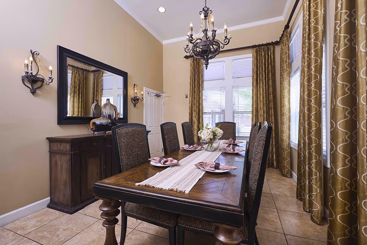 A formal dining room with a wooden dining table set for six, featuring pink napkins and a white table runner. The room has beige walls, large windows with patterned gold curtains, a dark wooden sideboard with decorative vases, a large rectangular mirror, and an ornate chandelier hanging from the ceiling.