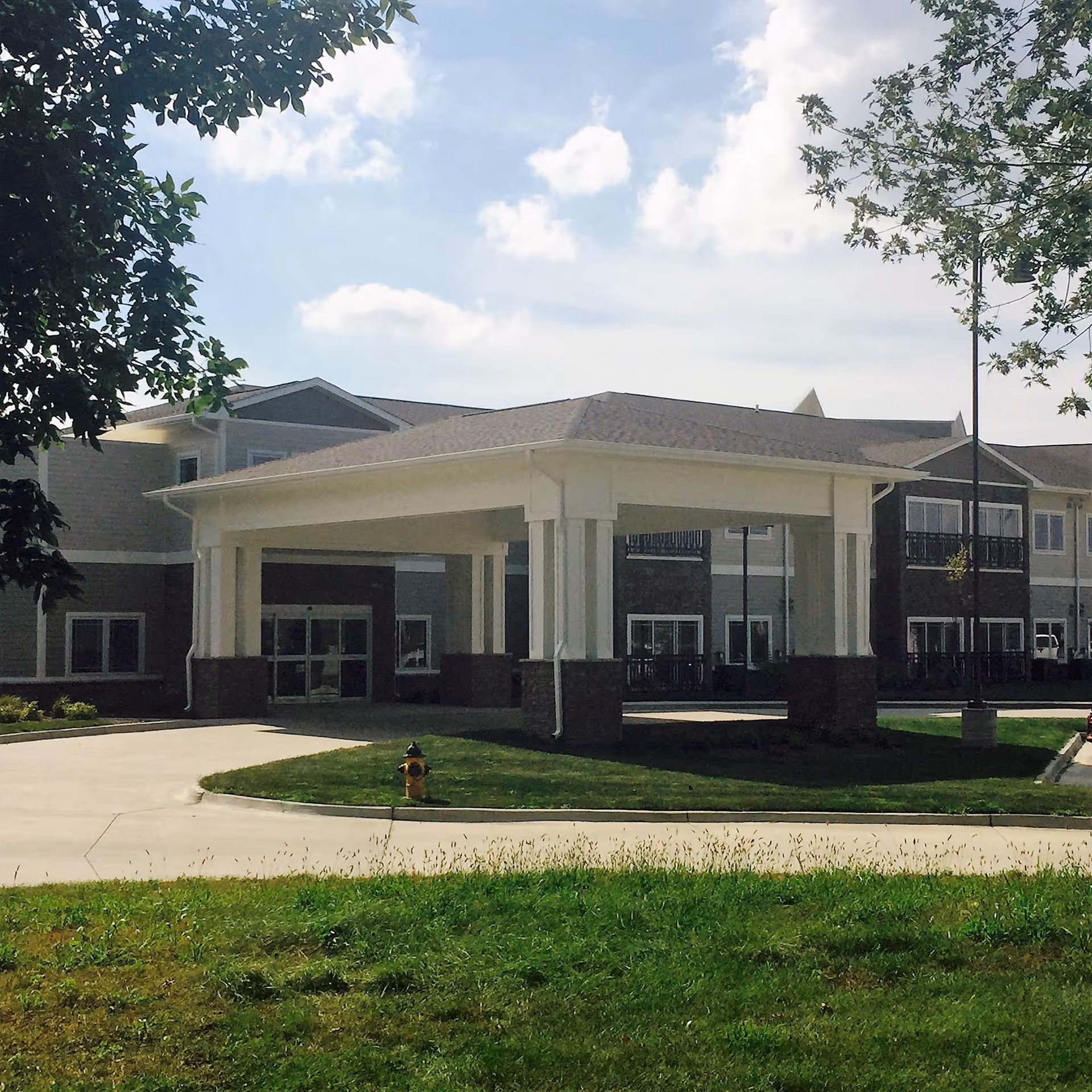 Exterior view of the Suites of Ankeny building showing the main entrance with a covered drop-off area, surrounded by green grass and trees under a partly cloudy sky.