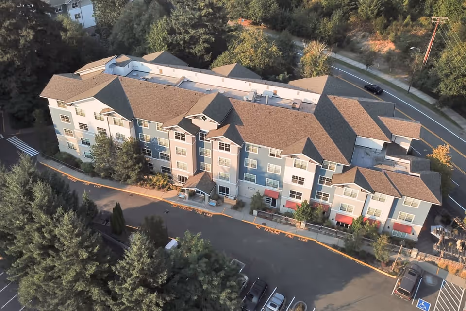 Aerial view of a multi-story senior living facility named The Creekside, surrounded by trees and adjacent to a road. The building has a complex roof structure and multiple windows, with a parking area in front including a handicapped parking space.