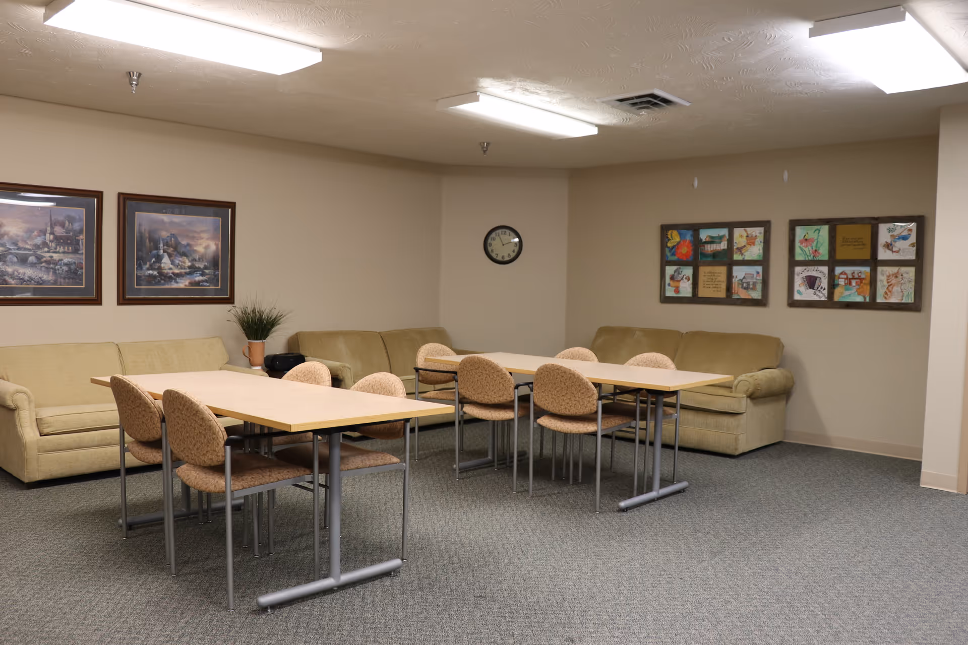 A common area in a residential care center with two beige sofas against the walls, two rectangular tables with eight cushioned chairs, framed artwork on the walls, a small potted plant, and a clock.