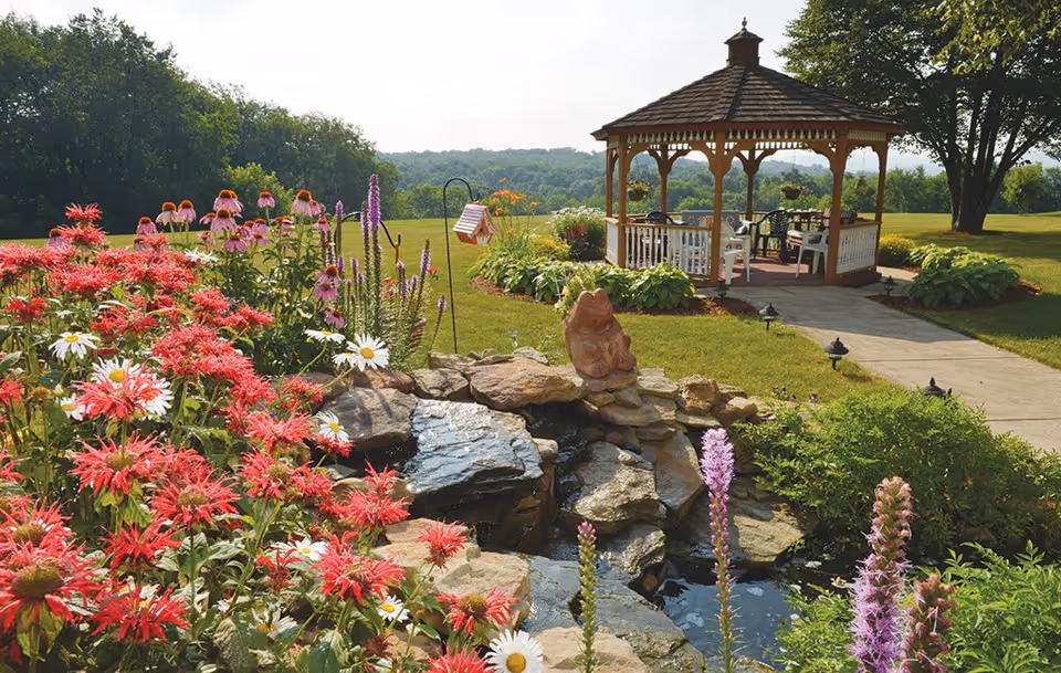 A landscaped outdoor garden with colorful flowers, a small stone pond, and a wooden gazebo overlooking a grassy lawn.