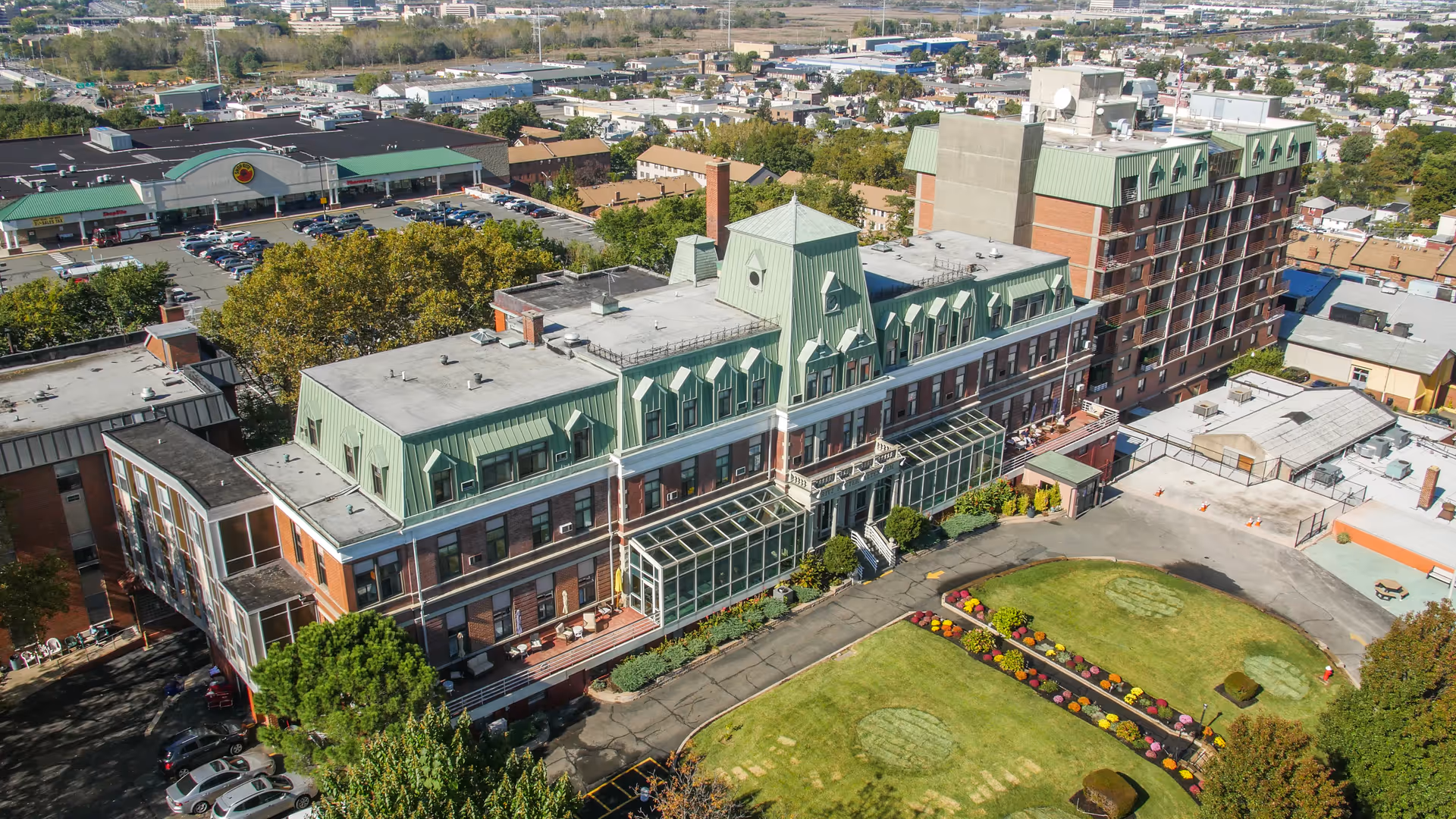Aerial view of a large senior living facility building with green roofs and multiple floors, surrounded by a well-maintained lawn with flower beds and a driveway. The building is located in an urban area with other buildings, parking lots, and streets visible in the background.