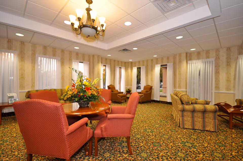 Bright senior living lounge with red upholstered chairs, floral centerpiece, and patterned carpet.