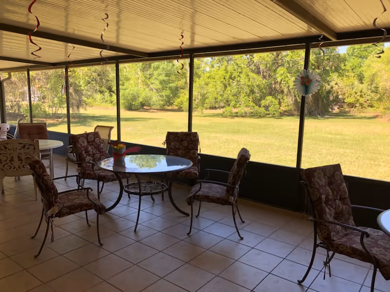 A covered patio area with several cushioned metal chairs arranged around glass-top tables. The patio has a tiled floor and is enclosed with large screened windows, offering a view of a grassy yard and trees outside. Some hanging decorations are visible from the ceiling.