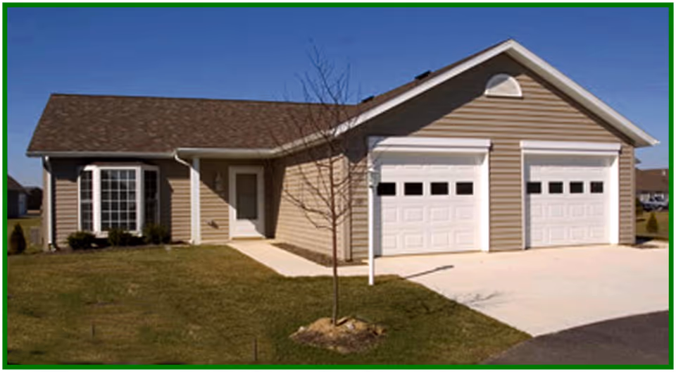 Single-story beige house with two white garage doors, a front door, bay window, driveway, and lawn with a small tree.