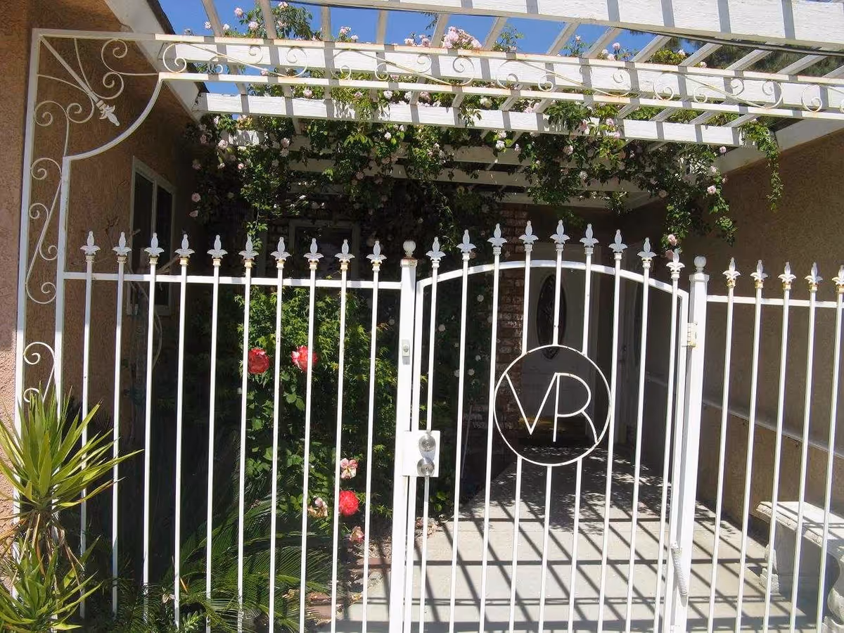 White wrought-iron gate with a circular monogram beneath a pergola and climbing roses at a home's entrance.