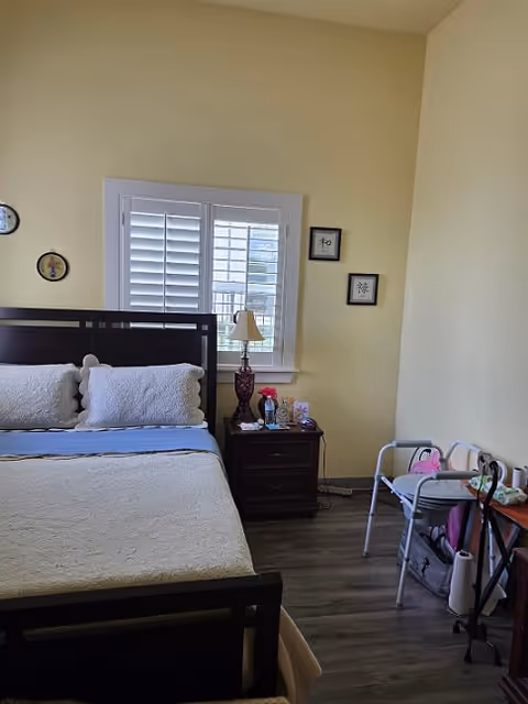 A bedroom with a dark wooden bed frame, white bedding, and two white pillows. There is a window with white shutters behind the bed. Next to the bed is a dark wooden nightstand with a decorative lamp, flowers, and some small items. On the right side of the room, there is a walker and a small table with various personal items. The walls are painted light yellow and the floor is dark wood.