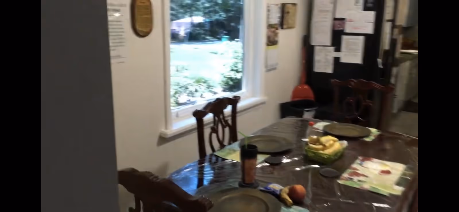 Dining area with a wooden table covered by a plastic tablecloth, set with placemats and plates. There is a basket of bananas and some fruit on the table. Wooden chairs surround the table. A window shows greenery outside, and a bulletin board with papers is visible in the background.