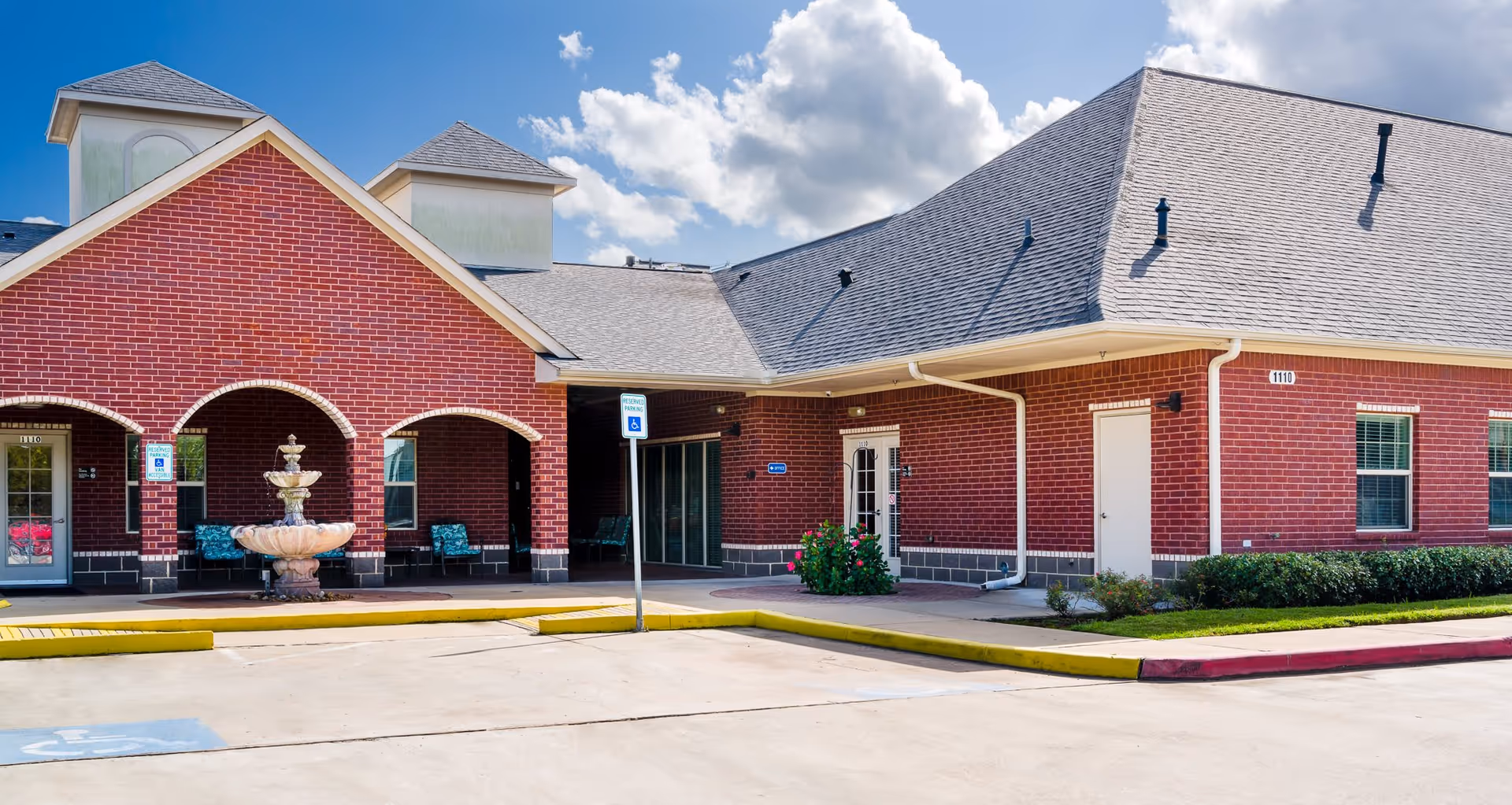 Front exterior of a red-brick senior living facility with an arched covered entrance, fountain, and adjacent parking area.