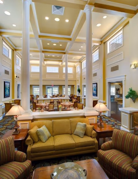Spacious, well-lit senior living dining and lounge area with tall columns, a coffered ceiling, sofa seating in the foreground and dining tables beyond.