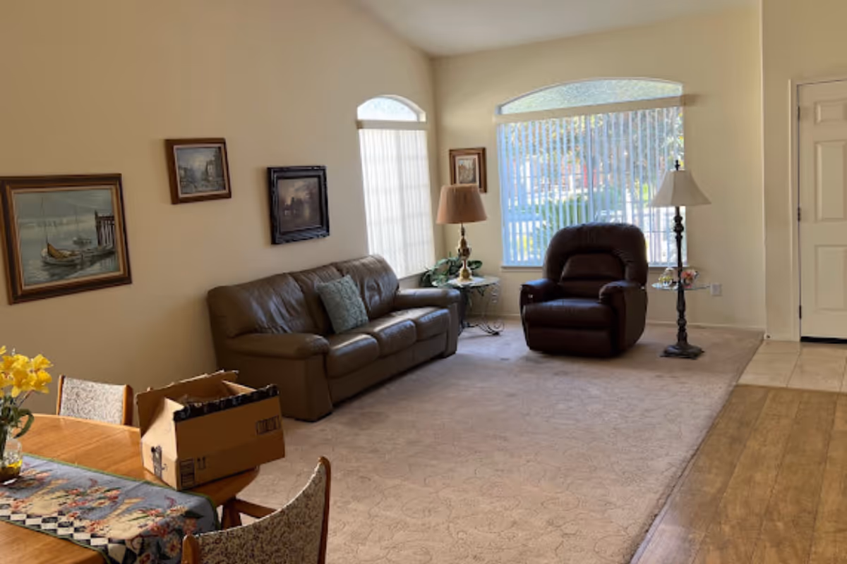 A cozy living room with a brown leather sofa, a dark brown recliner chair, two side tables with lamps, and three framed paintings on the wall. There is a large window with vertical blinds letting in natural light. A wooden dining table with chairs and a cardboard box is partially visible in the foreground.