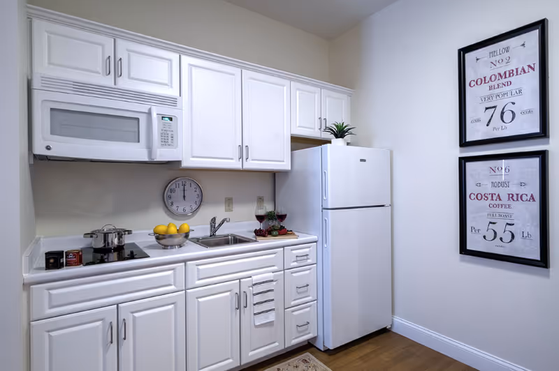 A small kitchen area with white cabinets, a white microwave, a white refrigerator, a sink, and a stovetop with a pot. There is a bowl of lemons on the counter, two glasses of red wine, and a small plant on top of the refrigerator. Two framed coffee-themed prints hang on the wall to the right.