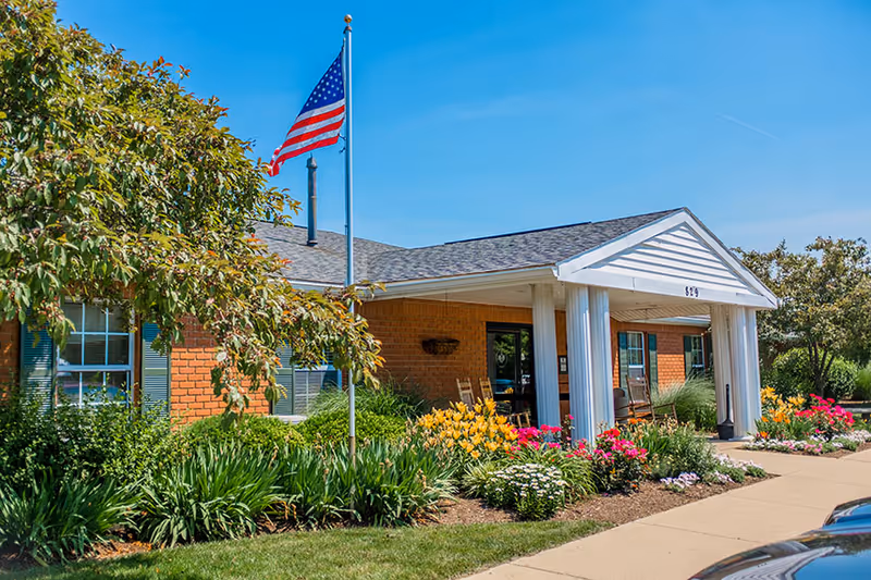 Front entrance of a brick senior living facility with a flagpole, white columns, rocking chairs, and colorful flower beds.