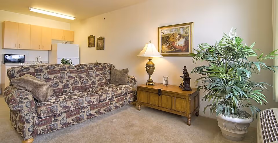 A cozy living room area featuring a patterned sofa with two cushions, a wooden side table with a decorative lamp and figurine, a large potted plant, and a framed painting of a dog on the wall. In the background, there is a small kitchen area with light wood cabinets, a white refrigerator, and a microwave.