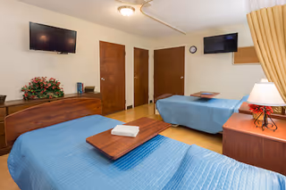 A room with two single beds covered with blue bedspreads, each with a wooden overbed table. There are two wooden dressers, one with a lamp and a small plant, and the other with a flower arrangement. Two flat-screen TVs are mounted on the walls. The room has three wooden doors and a clock on the wall.