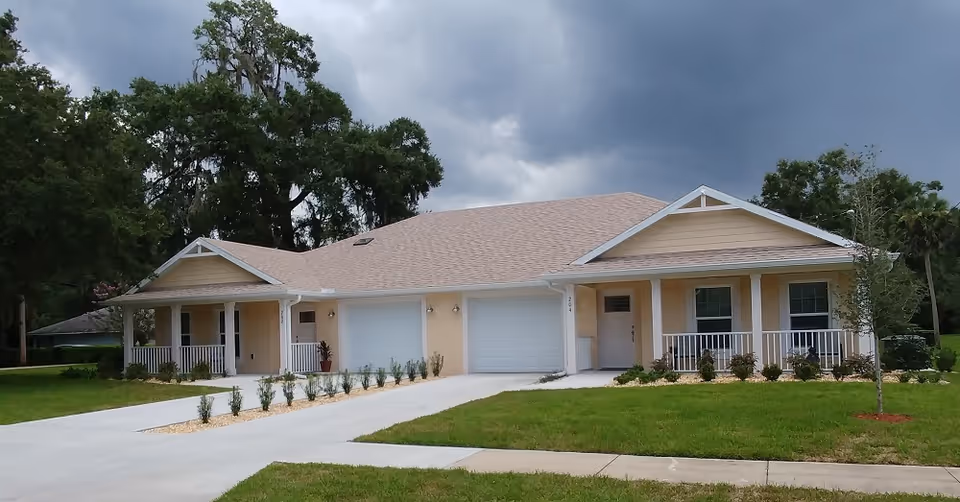 Exterior view of a single-story residential building with two attached garages, beige walls, and a light brown roof. The building has two covered porches with white railings and is surrounded by a green lawn and some small plants. The sky is cloudy and overcast.