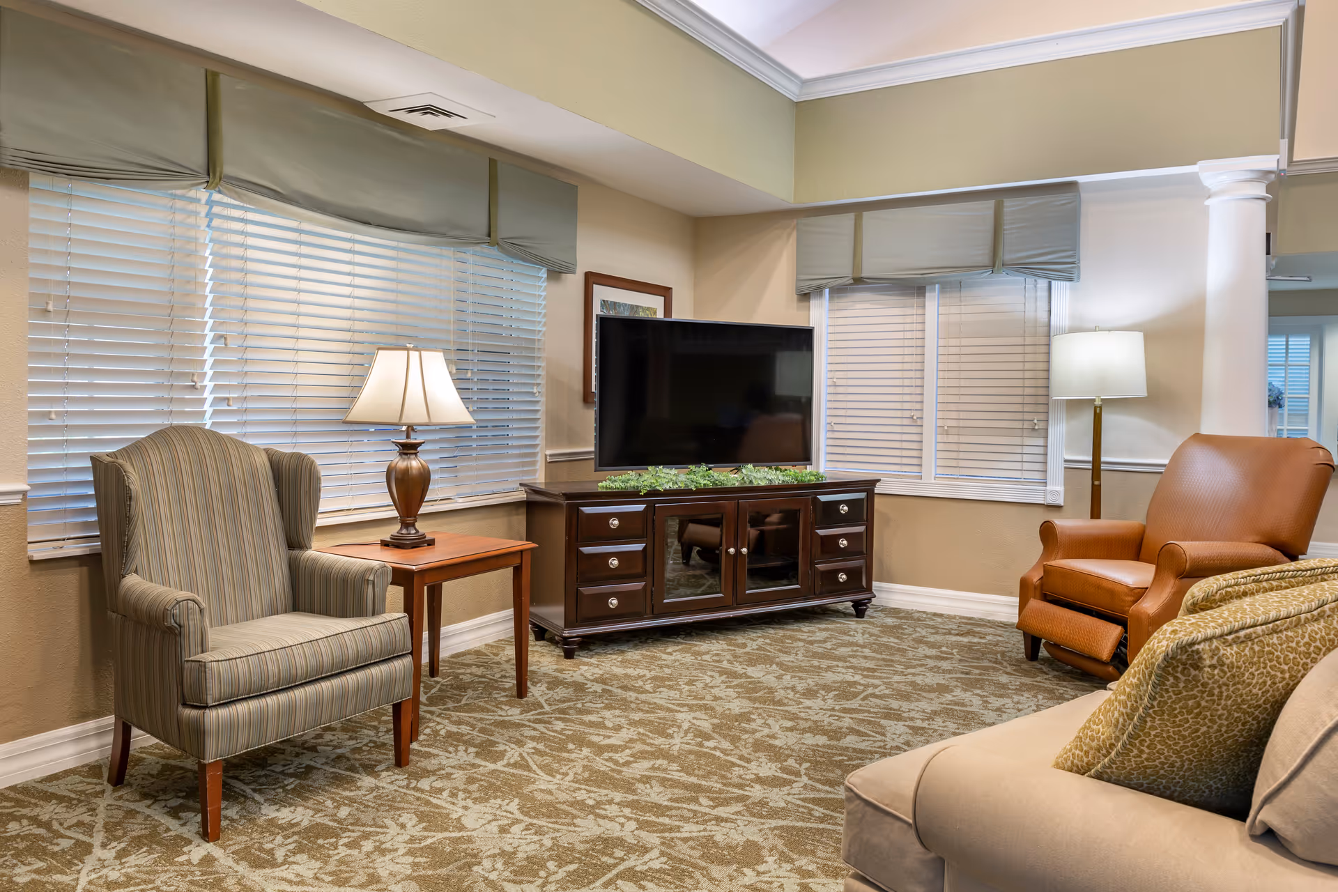 A cozy living room area with a beige sofa, a striped armchair, and a brown leather recliner. There is a wooden side table with a lamp between the sofa and armchair. A flat-screen TV sits on a dark wooden TV stand with drawers and cabinets. The room has large windows with white blinds and green valances, beige walls, and a patterned carpet.