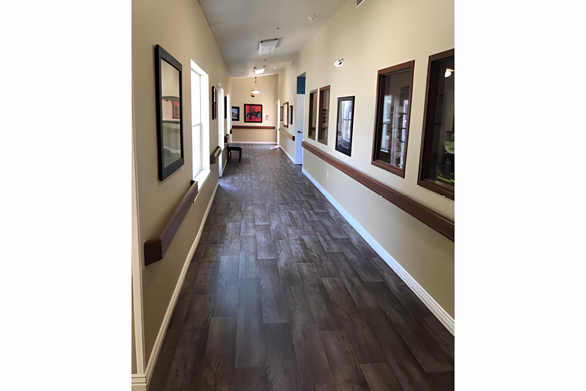 Long, well-lit interior hallway with wood-look flooring, handrails and framed artwork along the walls.