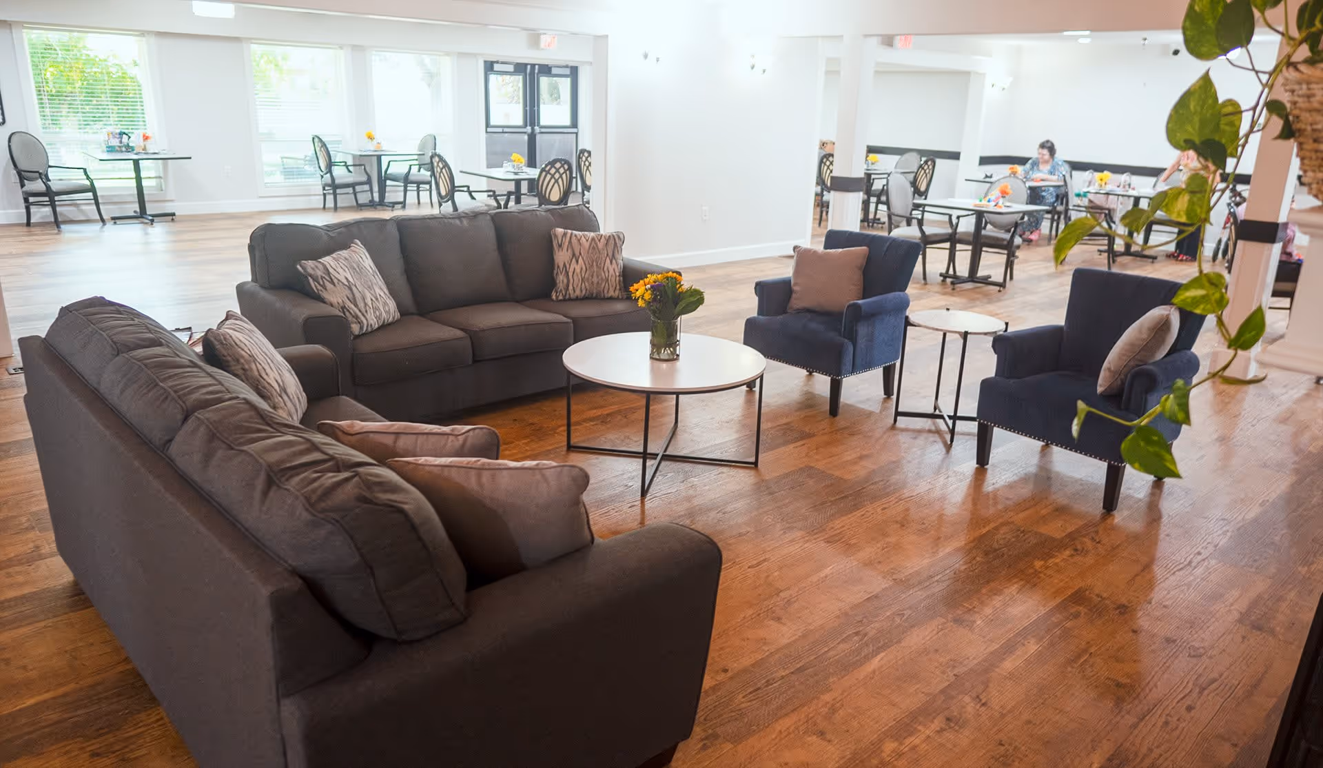 Spacious communal lounge with sofas, armchairs, coffee tables, and dining tables in a senior living facility.
