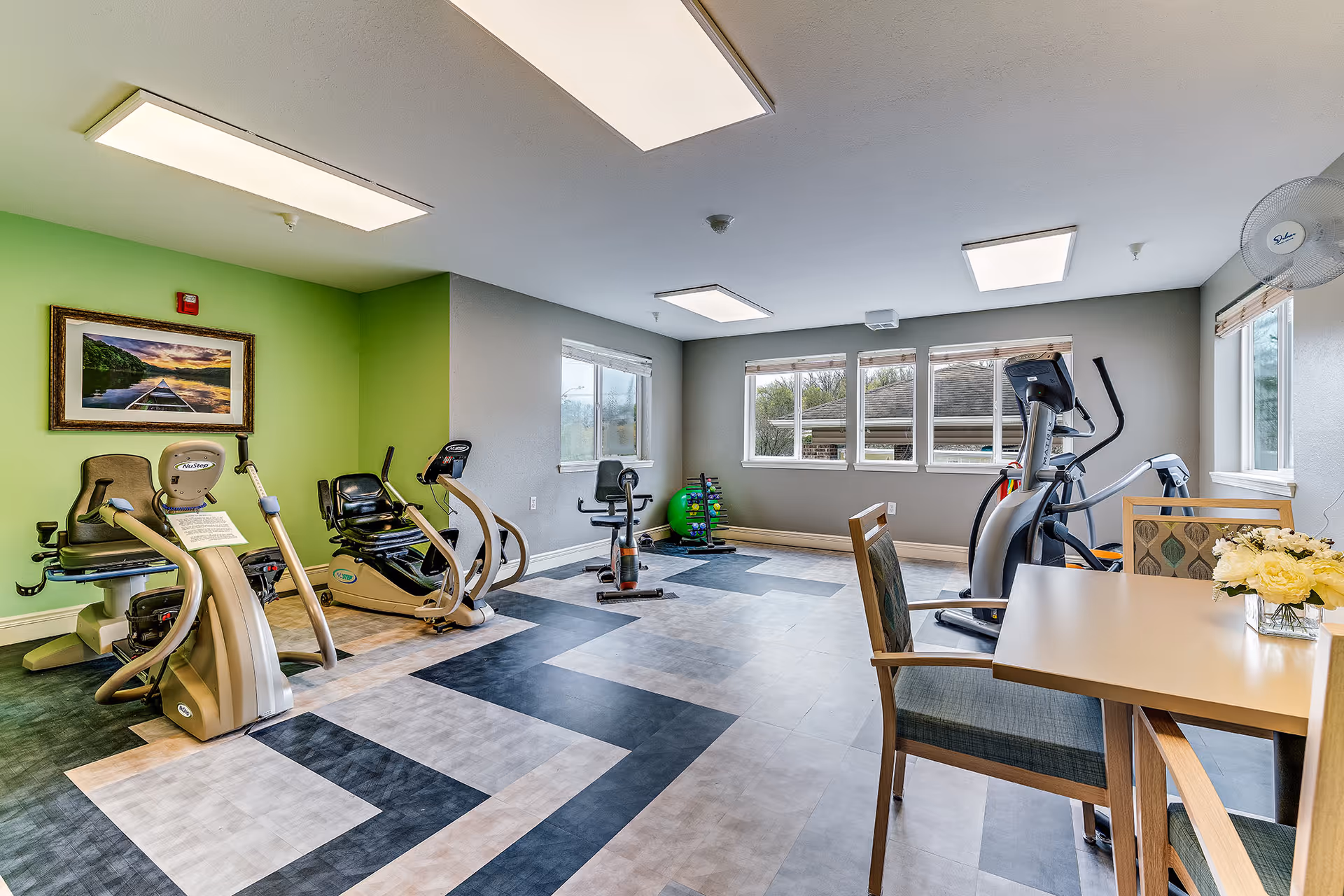 Bright interior exercise room with cardio machines, chairs and a table, patterned flooring and windows.