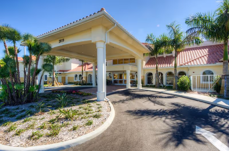 Entrance of a senior living facility with a covered drop-off area supported by white columns, surrounded by palm trees and landscaping under a clear blue sky.
