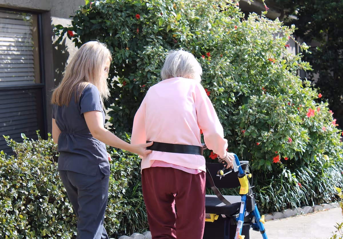 A caregiver assisting an elderly woman using a walker outside near greenery and flowers.