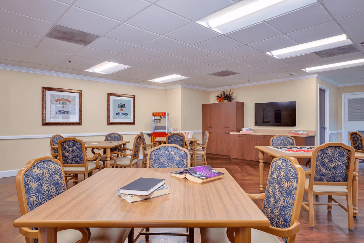 A common activity room in a senior living facility with several wooden tables and cushioned chairs. On the tables are books and a checkers game. The room has framed vintage car artwork on the walls, a popcorn machine in the corner, a flat-screen TV mounted on the wall, and a wooden cabinet with decorative plants on top.