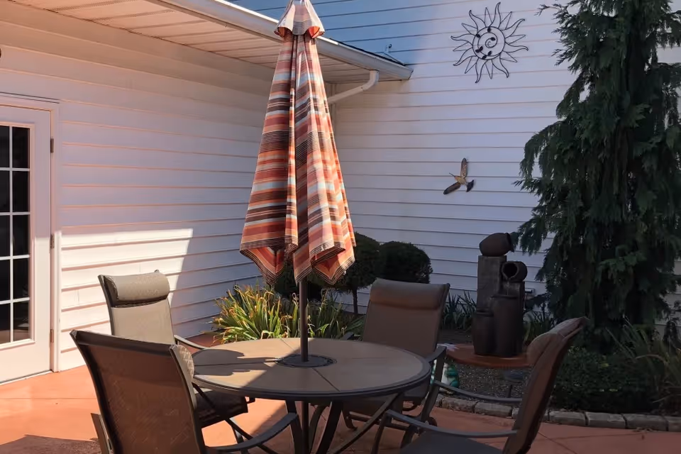 Outdoor patio area with a round table and four chairs. A closed striped umbrella is positioned in the center of the table. The patio is adjacent to a white building wall decorated with a metal sun and bird wall art. There are plants and a small sculpture near the wall.