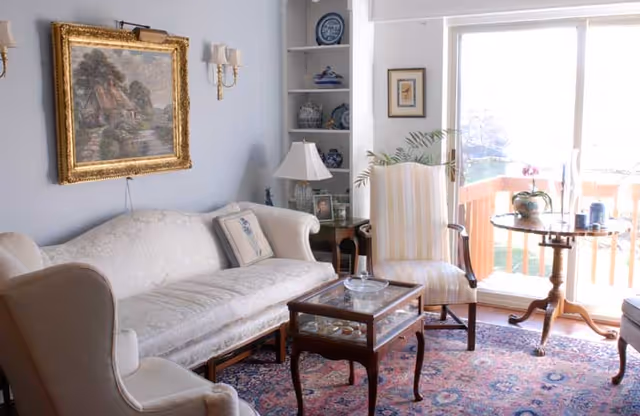 A cozy living room with a white upholstered sofa and armchair, a wooden coffee table with glass top, a striped armchair, and a round wooden side table near a sliding glass door leading to a balcony. The room features a patterned rug, a framed painting on the wall, built-in shelves with decorative items, and soft natural light coming through the door.