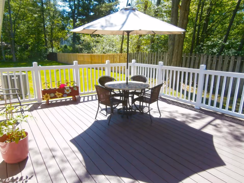 Outdoor patio area with a round table and four chairs under a large umbrella. The patio is surrounded by a white railing and there are potted plants and a flower box along the railing. Trees and a wooden fence are visible in the background.