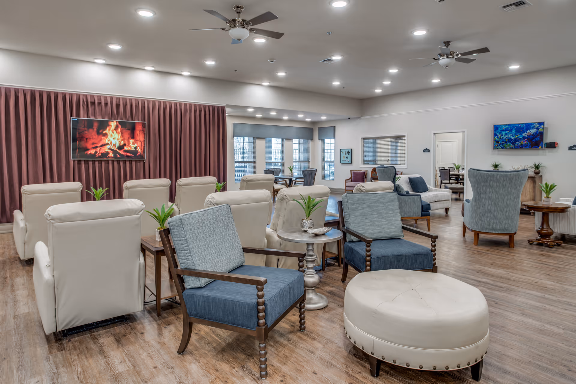 A spacious and well-lit common area in a senior living facility featuring multiple comfortable armchairs and small tables arranged on a wooden floor. Two flat-screen TVs are mounted on the walls, one displaying a fireplace and the other showing an underwater scene. Large windows with blinds allow natural light to enter the room, and ceiling fans are installed overhead.