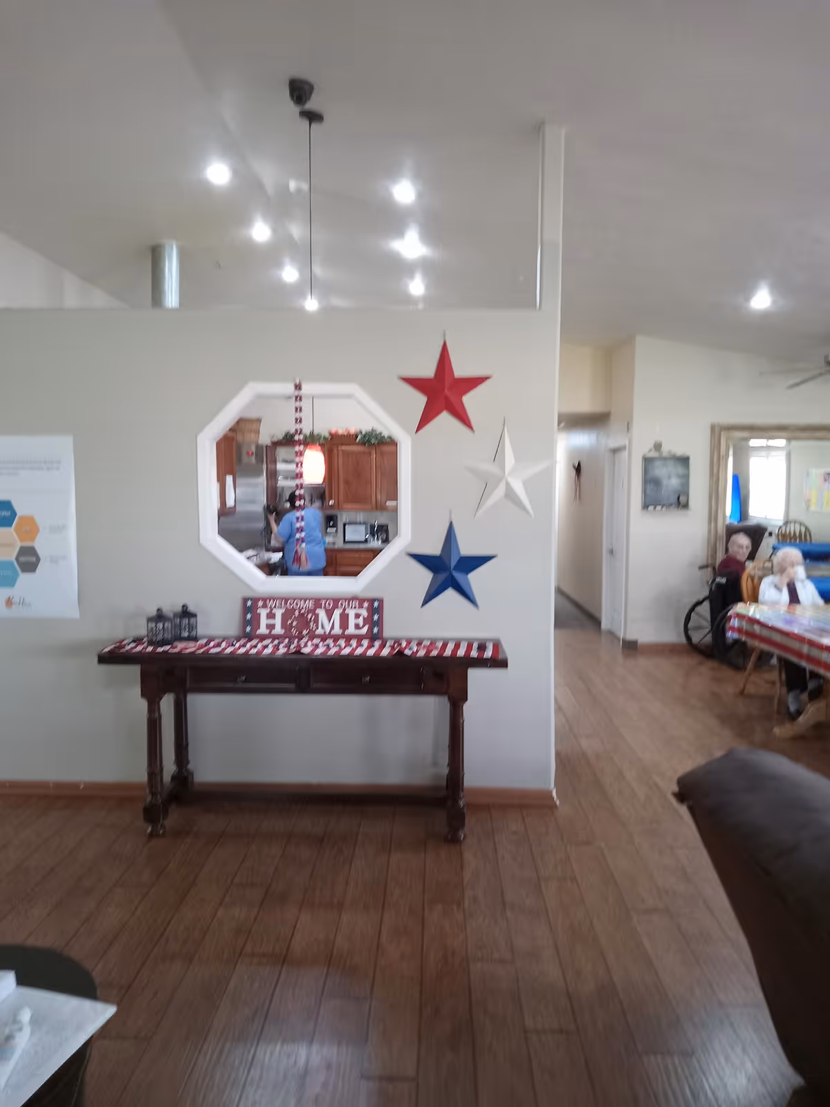 Interior common area of a senior living home featuring a console table under a wall decorated with red, white, and blue stars and a pass-through window to the kitchen, with a dining area and residents visible to the right.