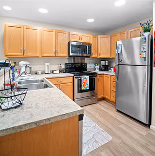 A modern kitchen with wooden cabinets, stainless steel appliances including a refrigerator, microwave, and stove. The countertop is light-colored with a double sink and various kitchen items such as a mixer, paper towel holder, and coffee maker. The floor has a light wood finish and there is a patterned rug in front of the stove.