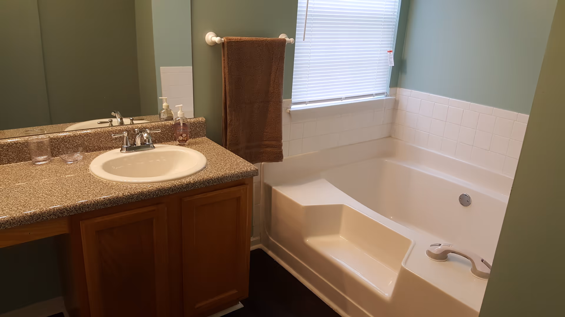 Bathroom with a white sink set in a speckled countertop with wooden cabinets below. A brown towel hangs on a white towel rack next to a white bathtub with built-in seating and a grab bar. A window with closed blinds is above the bathtub. The walls are painted light green and there is a large mirror above the sink.