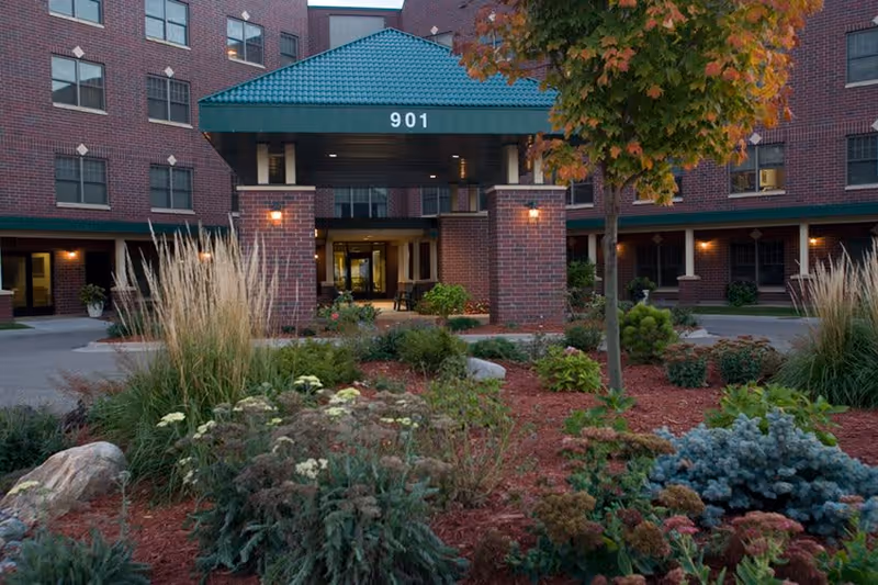 Entrance of a senior living facility named The Glenn Hopkins with a covered driveway, brick exterior, and landscaped garden with various plants and shrubs in the foreground.