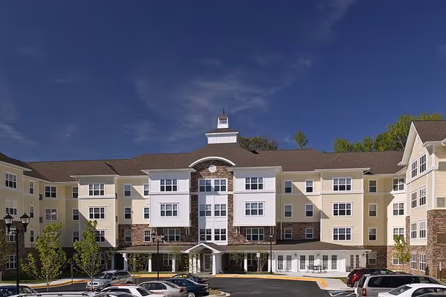 Exterior view of a large, multi-story senior living facility building with beige and white siding, stone accents, multiple windows, and a cupola on the roof. Several cars are parked in front of the building under a clear blue sky.
