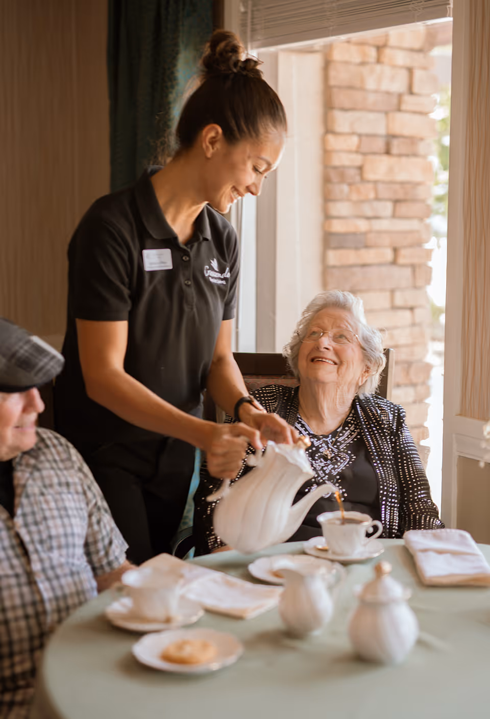 A caregiver in a black polo shirt is pouring tea from a white teapot into a teacup for an elderly woman who is smiling and looking up at her. They are seated at a round table with a light green tablecloth, white napkins, and a plate with a cookie. Another elderly person is partially visible sitting next to the woman. The setting appears to be a cozy indoor dining area with a window and stone wall in the background.
