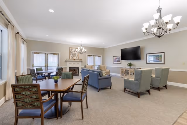 A spacious senior living common area with multiple seating options including a wooden table with four chairs, a blue sofa, and two armchairs. The room features a fireplace with a decorative painting above it, a large flat-screen TV mounted on the wall, and two framed pictures. Large windows with curtains allow natural light to fill the room, and two chandeliers provide additional lighting.