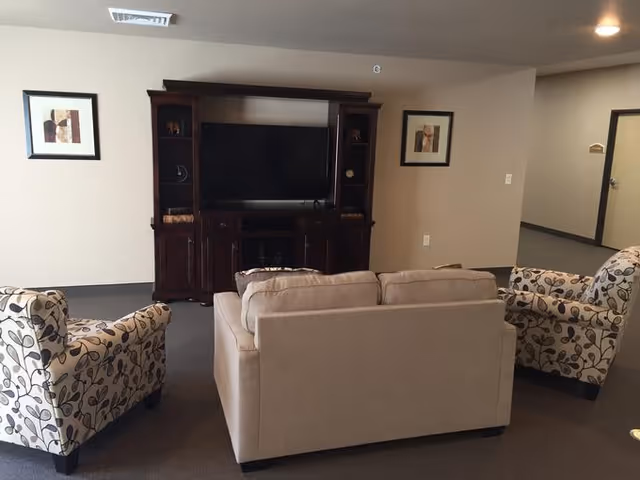 A cozy living room area with a beige sofa and two patterned armchairs facing a dark wooden entertainment center with a flat-screen TV. The walls are light-colored with two framed abstract artworks on either side of the TV. The room has carpeted flooring and a hallway visible in the background.