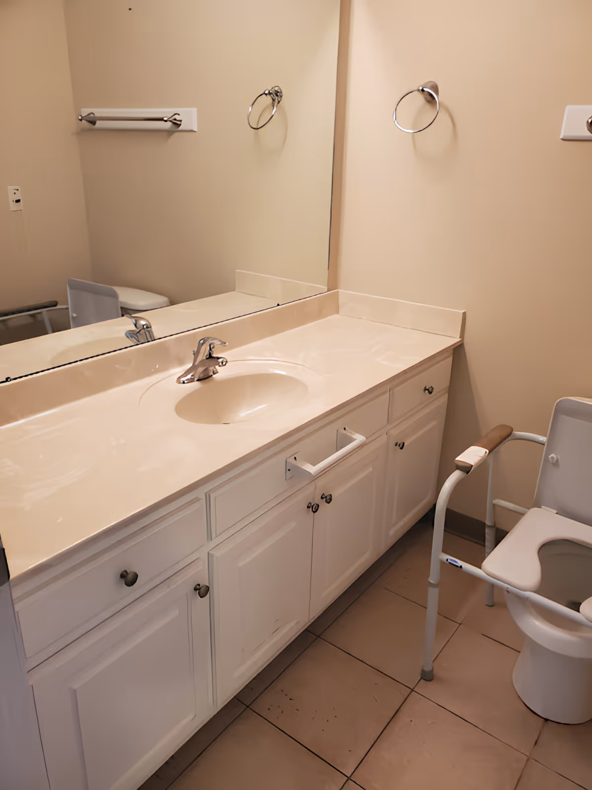 Bathroom with a beige countertop and sink, a large mirror above the sink, white cabinets below, a towel ring on the wall, and a raised toilet seat with armrests.